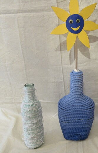 Zwei Flaschen, eine ist mit weißer Kordel umwickelt, die andere mit blauer Wolle. In der blauen Flasche steckt eine gelbe Papierblume